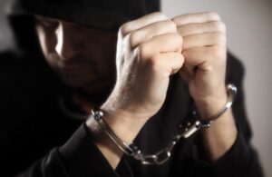 A dark photo of a man in handcuffs with his hands obscuring his face.