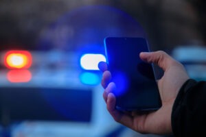 Officer holding a smartphone with a cop car in the background after asking to search someone’s phone.